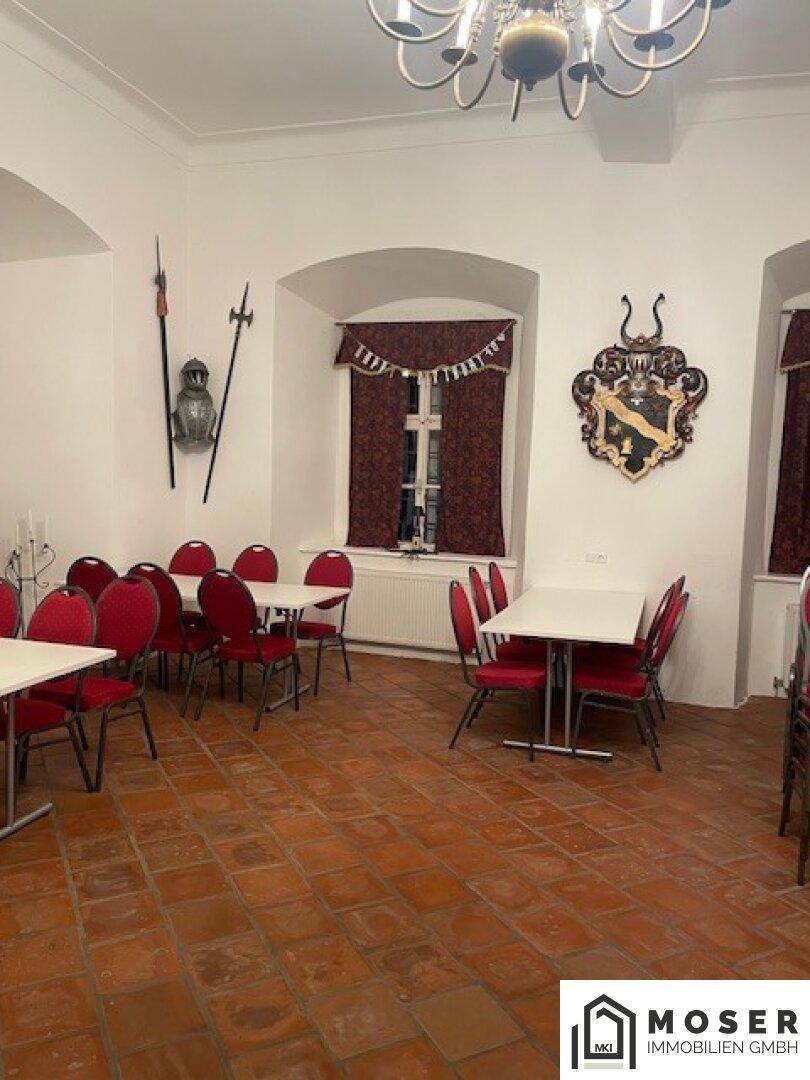 Veranstaltungsraum mit Terrakotta-Boden, roten Stühlen und Tischen, dekoriert mit historischen Wappen und Waffen.