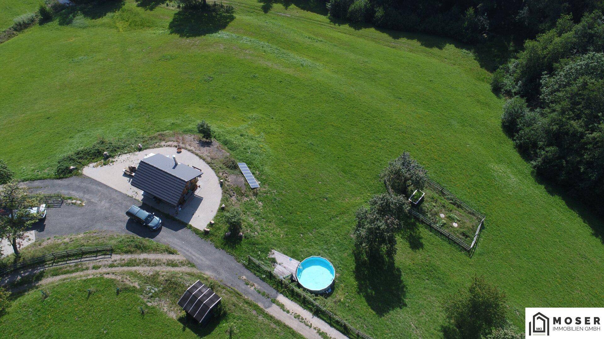 Luftaufnahme der Hütte mit Pool, Solarpaneelen und umzäuntem Gemüsegarten, umgeben von grünen Wiesen.