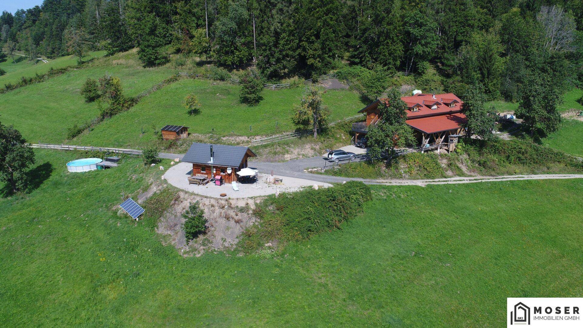 Nähere Luftaufnahme des Anwesens mit Haupthaus, Holzhütte, kleinem Pool und Solarpaneelen in grüner Umgebung.