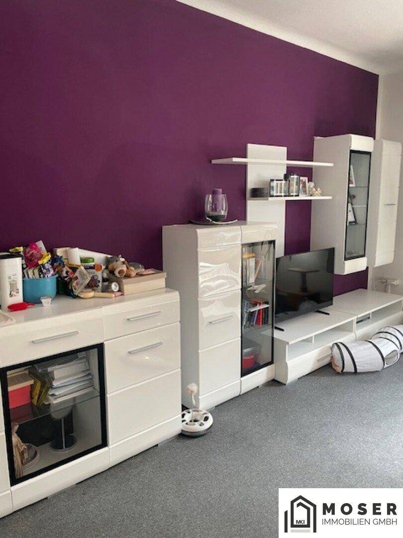 Wohnzimmer mit moderner weißer Schrankwand, lila Wandfarbe und Teppichboden.