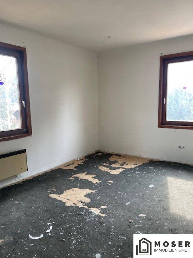 Leeres Zimmer mit zwei Fenstern und stark beschädigtem Bodenbelag.