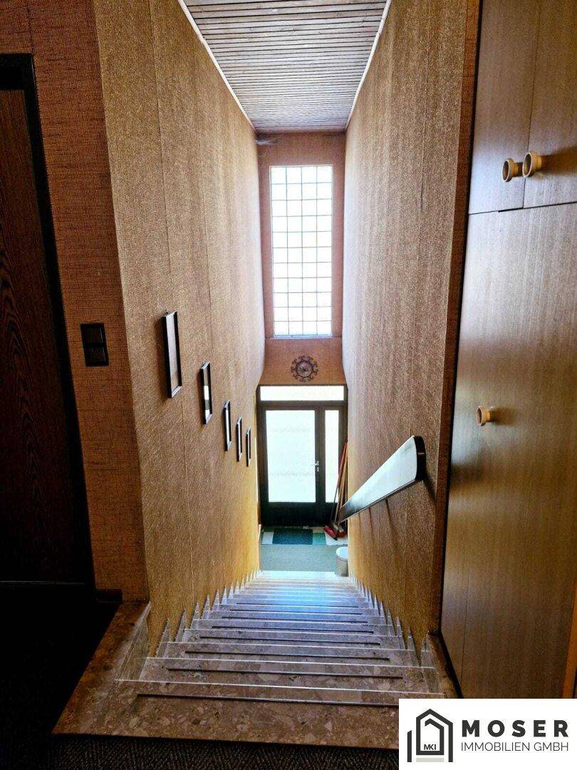 Treppenhaus mit Marmorstufen, Holzvertäfelung und einem hohen Fenster mit Glasbausteinen.