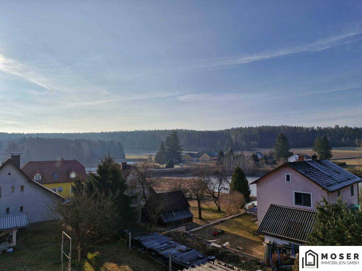 Weitläufiger Blick über eine ländliche Landschaft mit Häusern, Wald und einem See im Hintergrund.