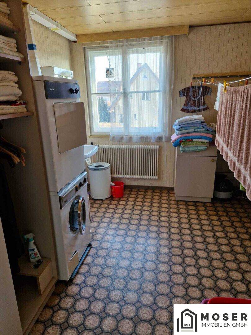 Waschküche mit Waschmaschine, Trockner, Regalen und einem Fenster mit Blick ins Grüne.