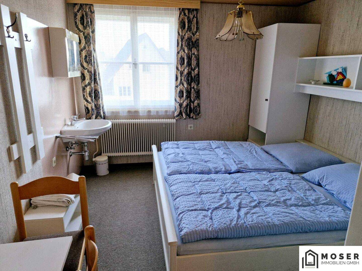Schlafzimmer mit Doppelbett, Waschbecken und Kleiderschrank, hell durch großes Fenster.