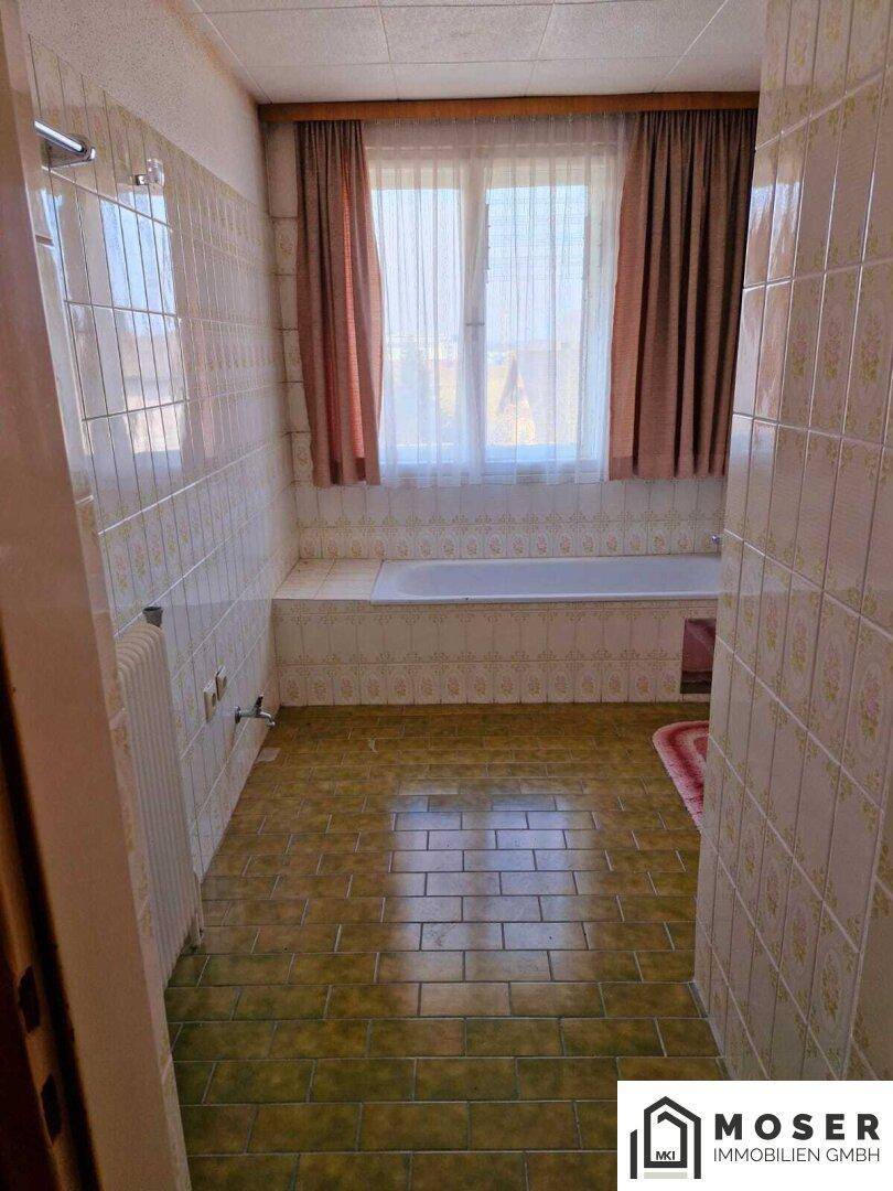 Badezimmer mit Badewanne, Fliesenboden und einem Fenster mit braunen Vorhängen.