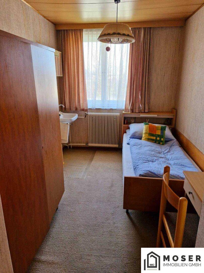 Einzelzimmer mit Bett, Kleiderschrank, Waschbecken und einem Fenster mit Vorhängen.