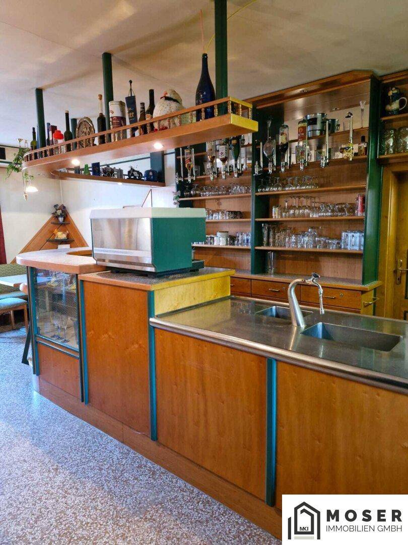 Bar-Theke mit Spüle, Kaffeemaschine und Regalen voller Flaschen und Gläser.