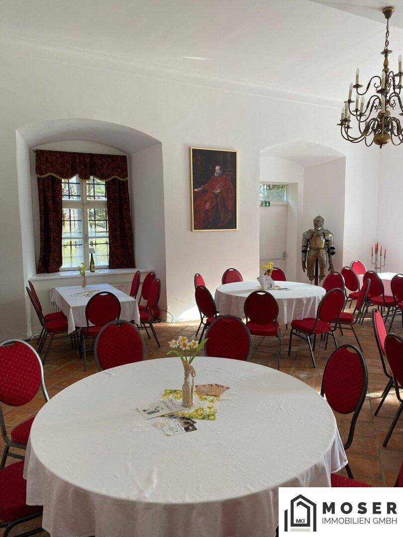 Heller Veranstaltungsraum mit festlich gedeckten Tischen, roten Stühlen und historischen Gemälden an den Wänden.