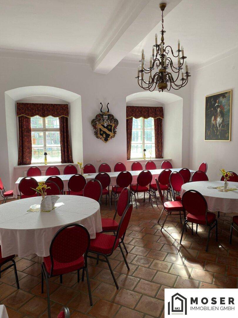 Großer Veranstaltungsraum mit Terrakotta-Fliesenboden, runden Tischen und roten Stühlen, beleuchtet von einem Kronleuchter.
