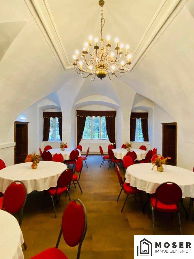 Elegante Veranstaltungshalle mit gewölbter Decke, runden Tischen und roten Stühlen, beleuchtet von einem großen Kronleuchter.