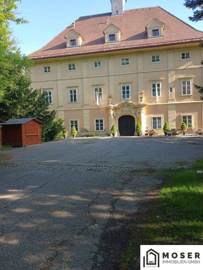 Ansicht des historischen Gebäudes mit Zufahrt und umgebenden Bäumen an einem sonnigen Tag.