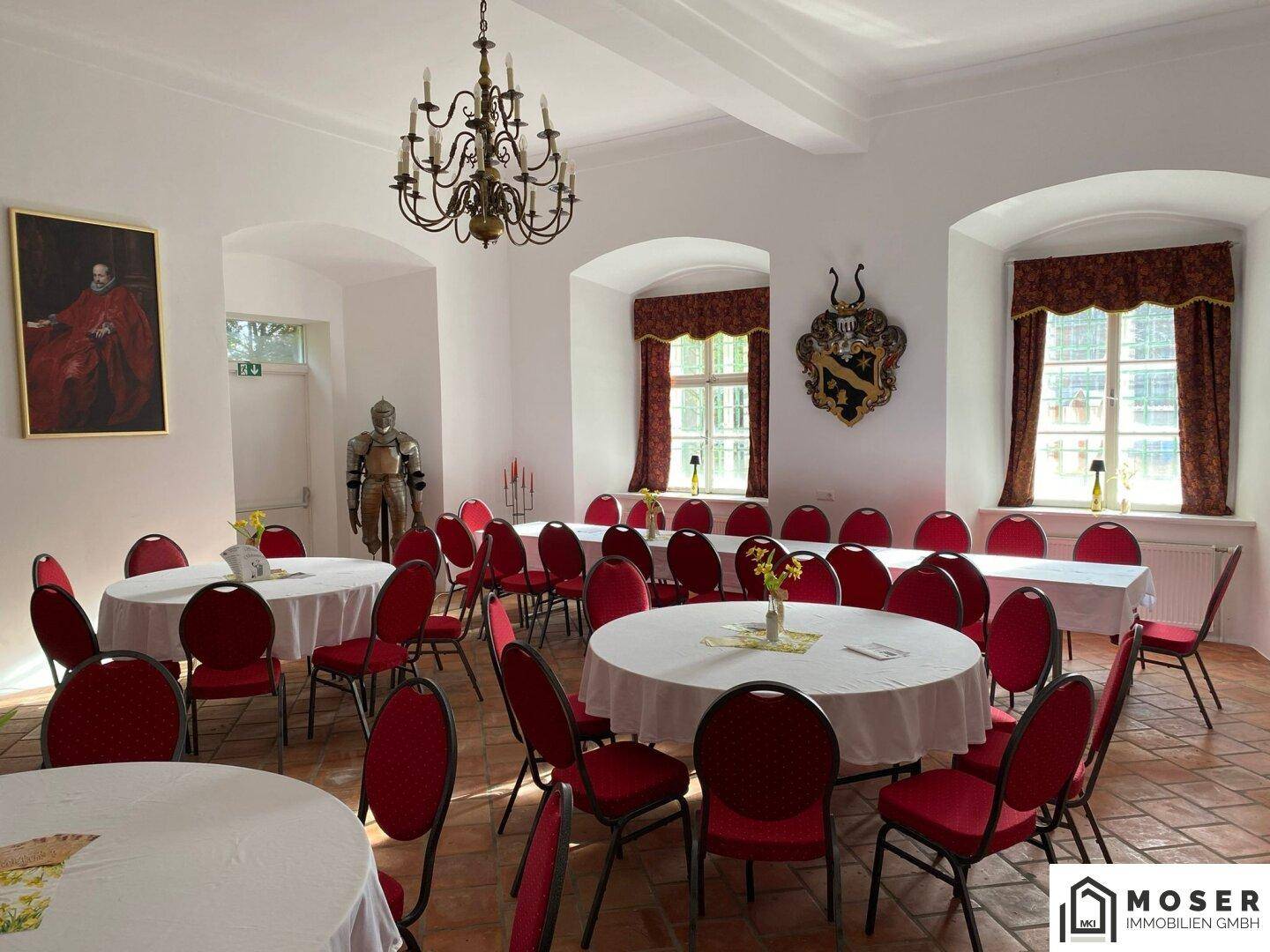 Großer, heller Veranstaltungsraum mit verschiedenen Tischarrangements, roten Stühlen und historischen Dekorationen.