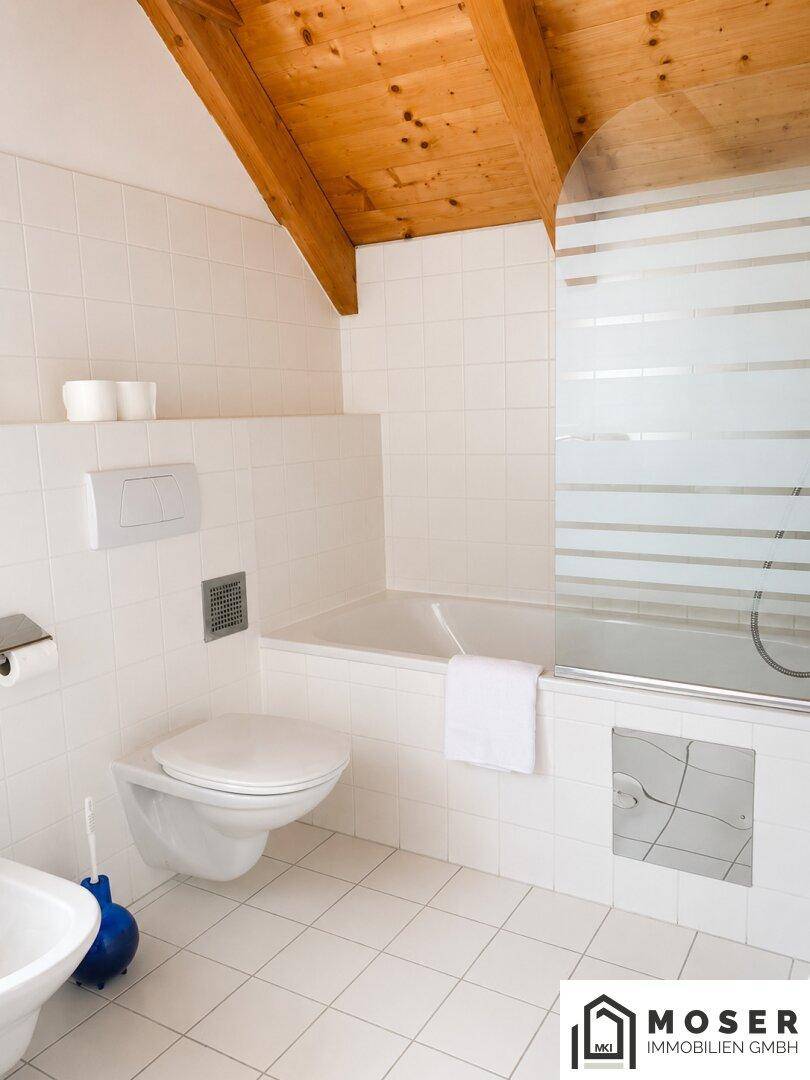 Modernes Badezimmer mit Badewanne, WC, weißen Fliesen und einer charmanten Holzdecke mit Dachschräge.