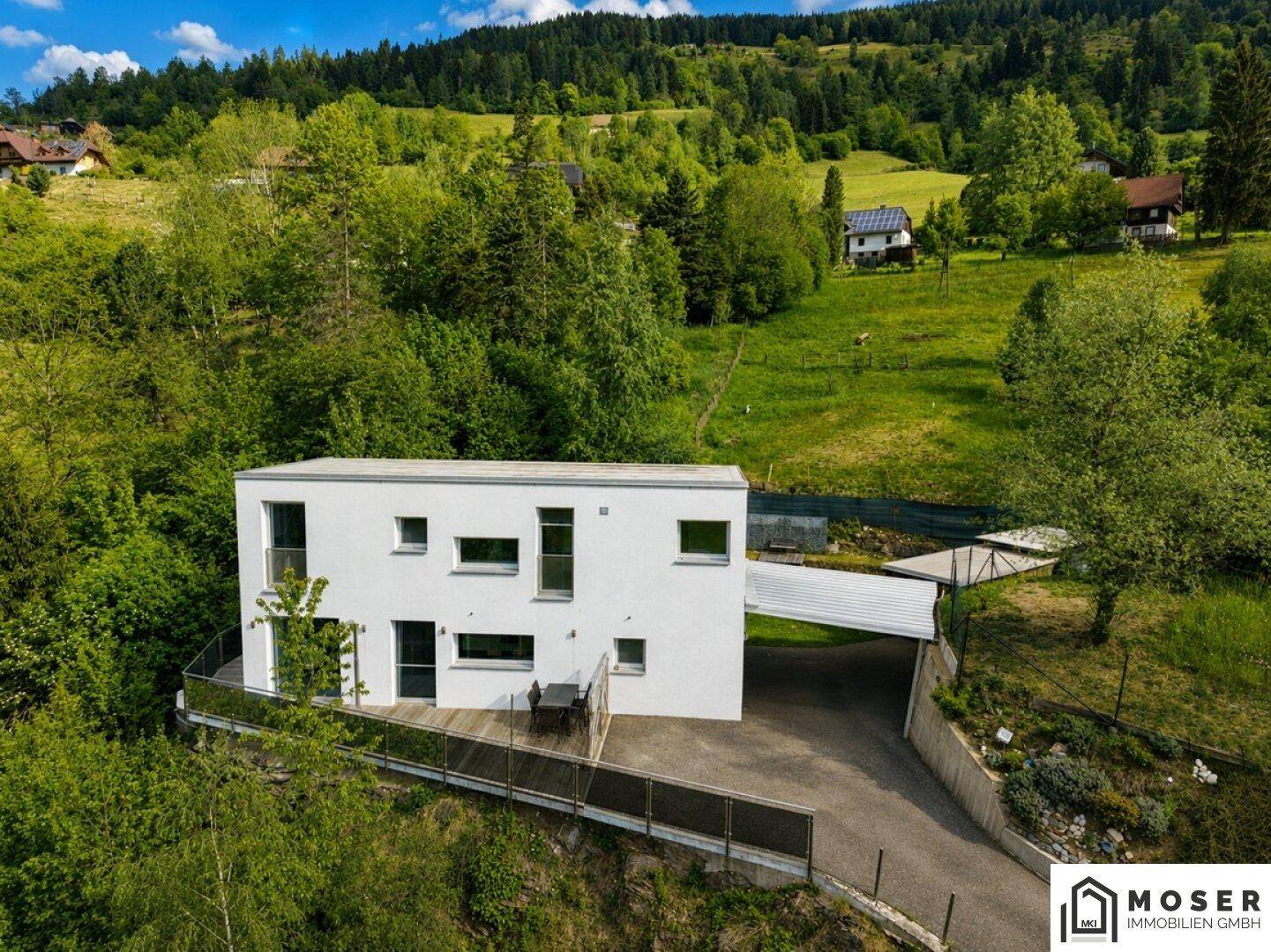 Ansicht des modernen Hauses mit Carport und Zufahrt, umgeben von grüner Landschaft.
