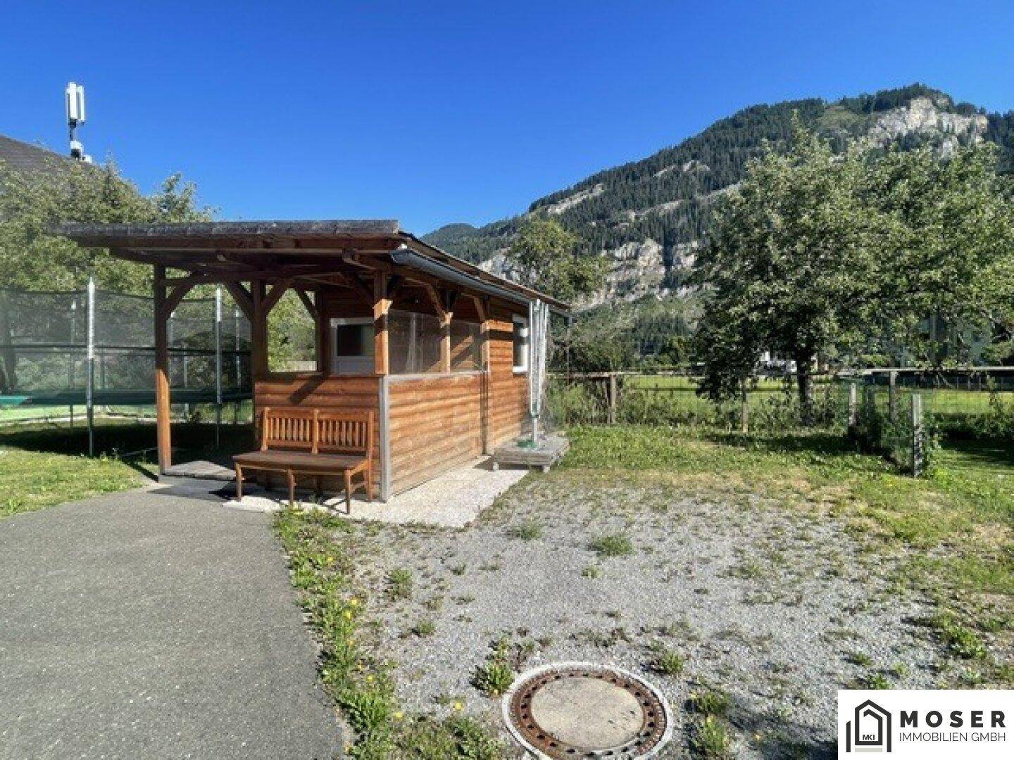 Idyllischer Gartenbereich mit einem rustikalen Holzhaus und Sitzbank unter blauem Himmel.