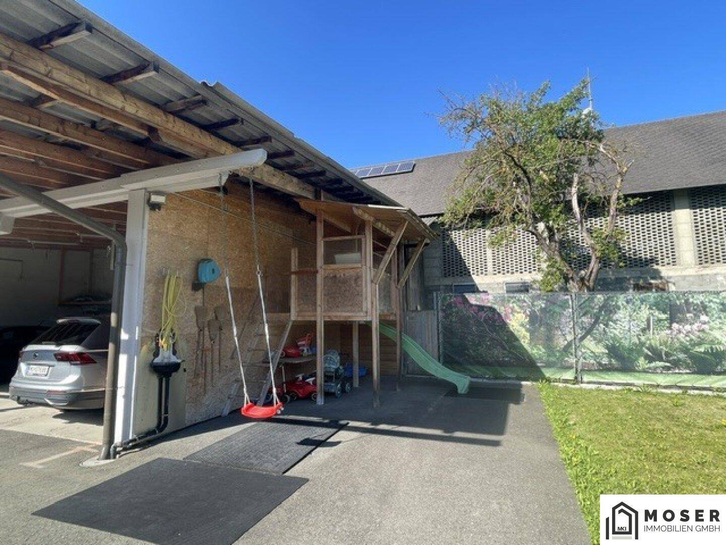 Spielbereich im Garten mit Rutsche und Schaukeln neben dem Carport, umgeben von Grün.