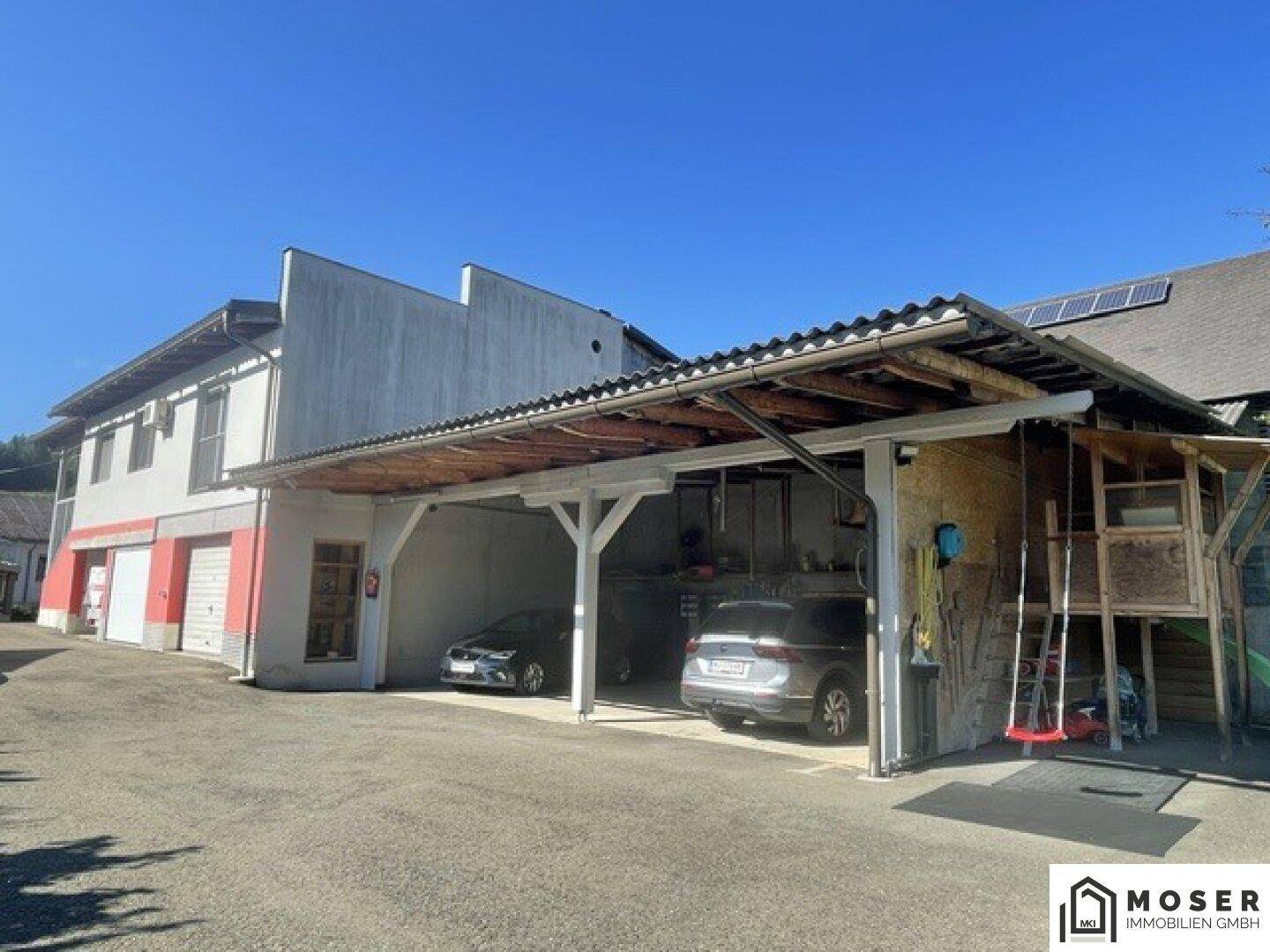 Seitenansicht des Gebäudes mit überdachtem Carport und geparkten Fahrzeugen an einem sonnigen Tag.
