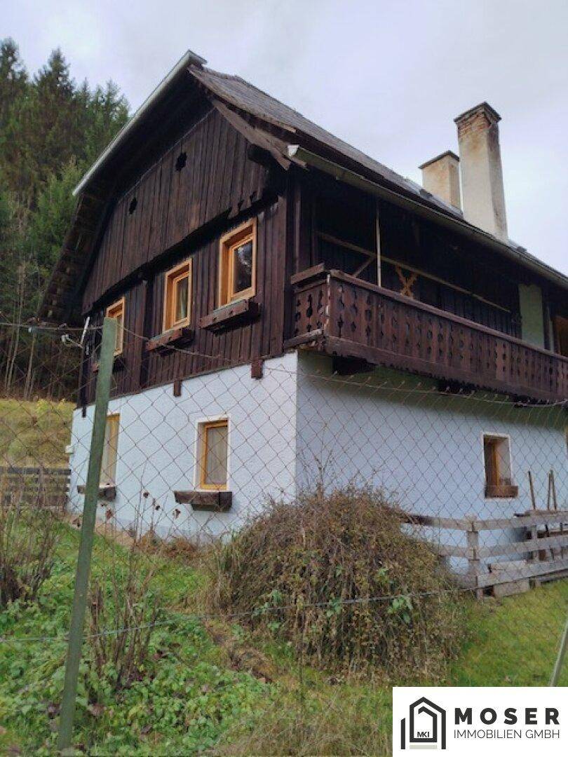 Traditionelles Haus mit Holzbalkon und Satteldach vor bewaldeter Bergkulisse.