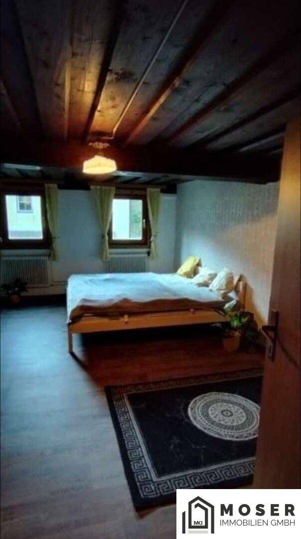 Schlafzimmer mit Holzboden, Holzbalkendecke und zwei Fenstern für natürliches Licht.