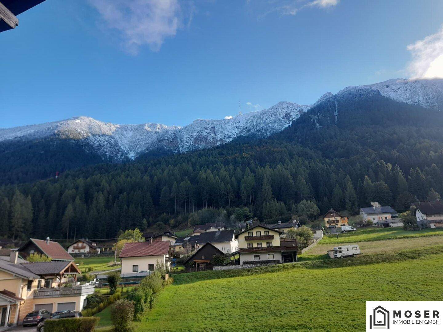 Panoramablick auf schneebedeckte Berge und grüne Hügel mit verstreuten Häusern unter blauem Himmel.