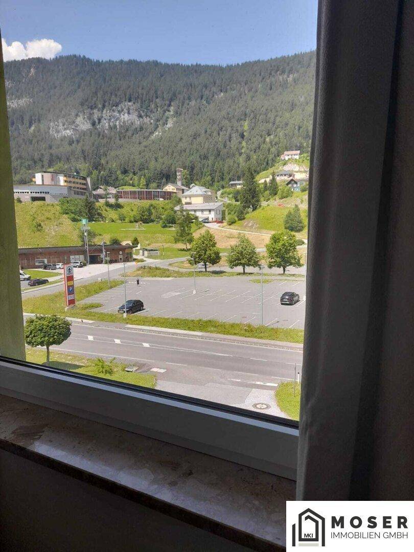 Blick aus dem Fenster auf eine Straße, Parkplätze und bewaldete Berge im Hintergrund.