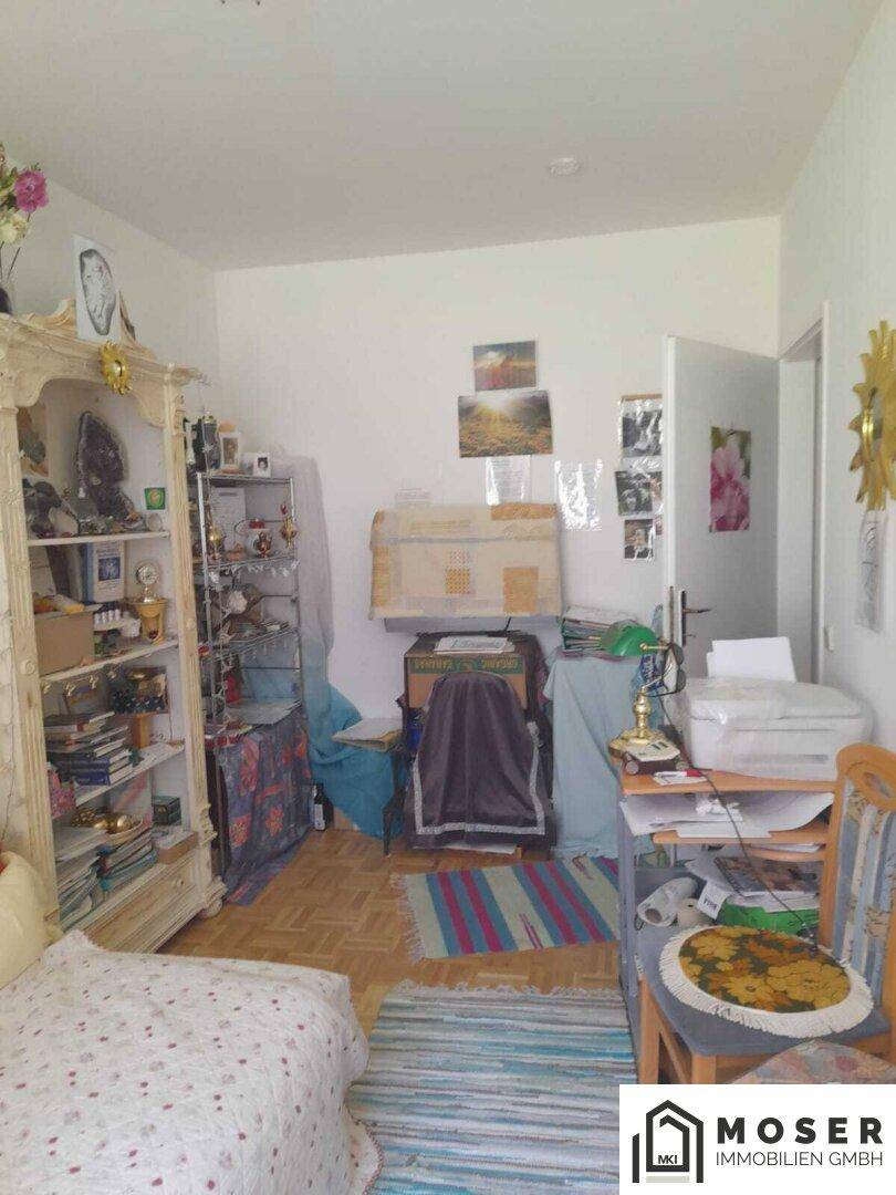 Helles Zimmer mit Holzparkett, Bücherregal und Schreibtisch, das als Schlaf- oder Arbeitszimmer dient.