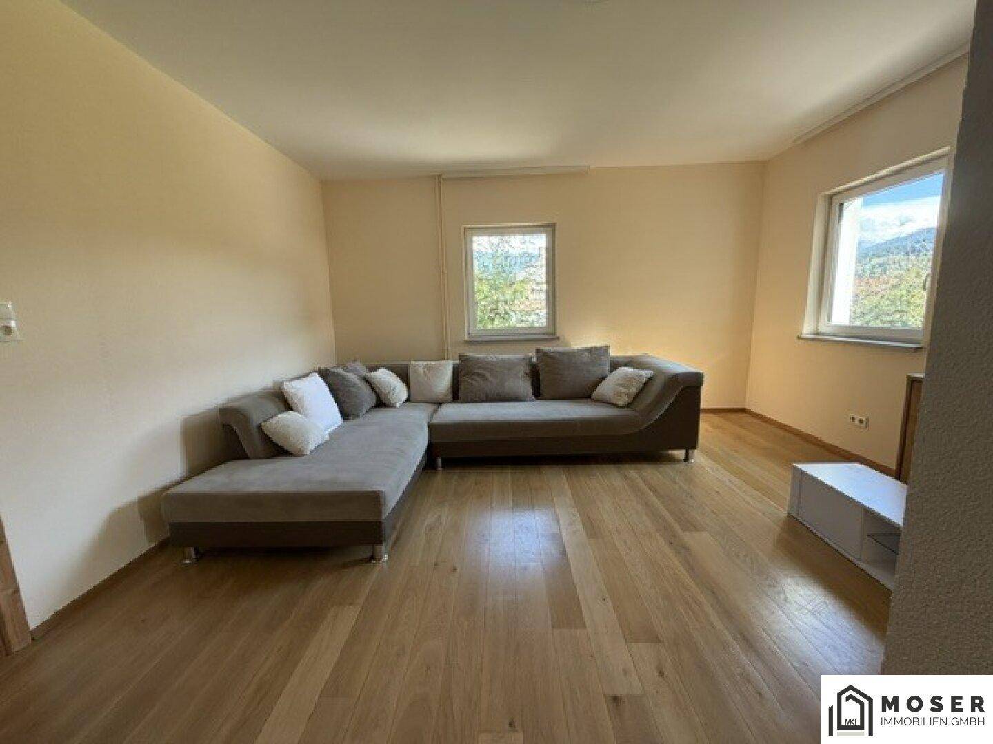 Helles Wohnzimmer mit großem Ecksofa, Holzboden und zwei Fenstern mit Ausblick ins Grüne.