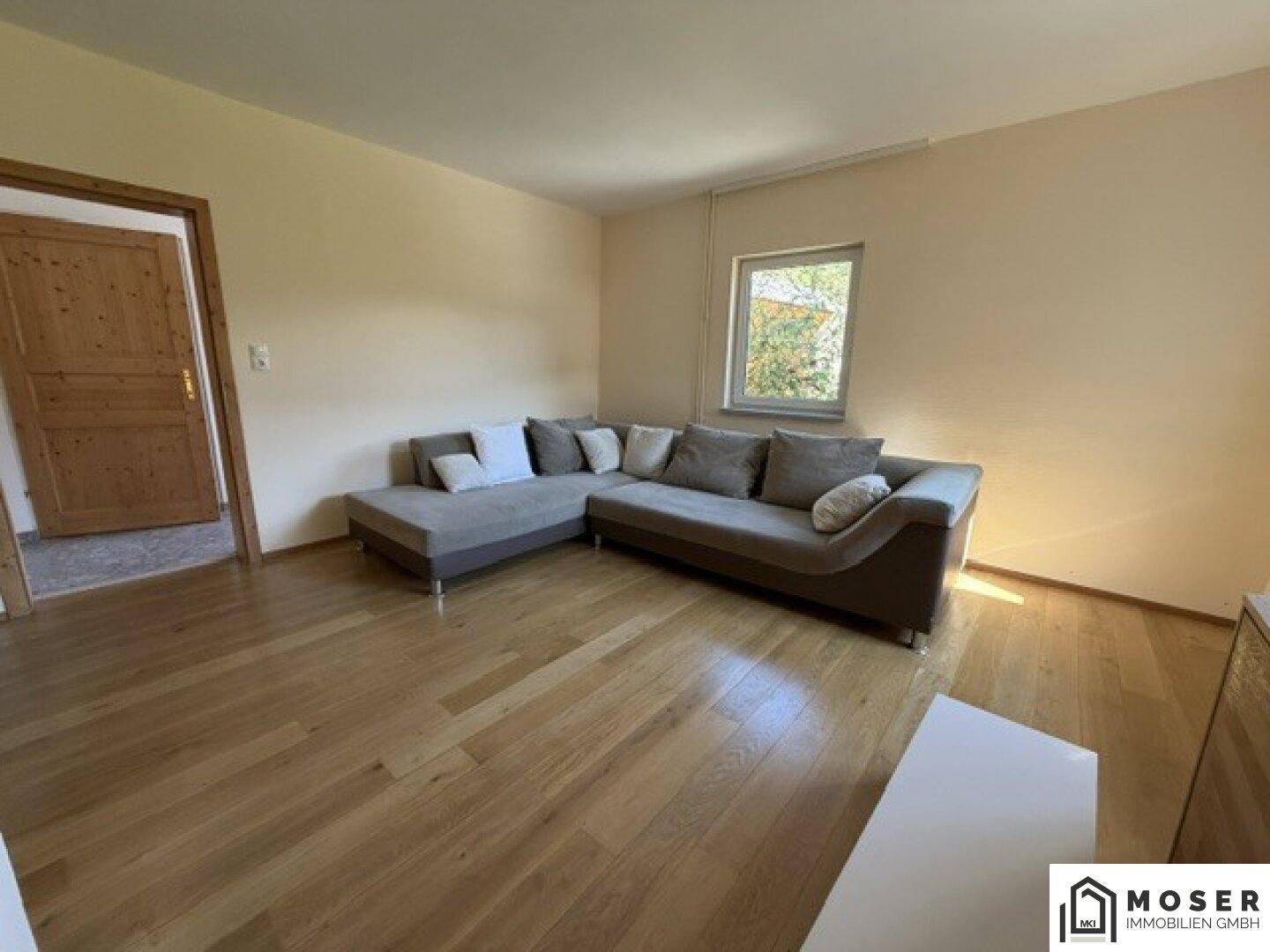 Geräumiges Wohnzimmer mit Ecksofa, Holzboden und Zugang zu weiteren Räumen, hell und einladend.