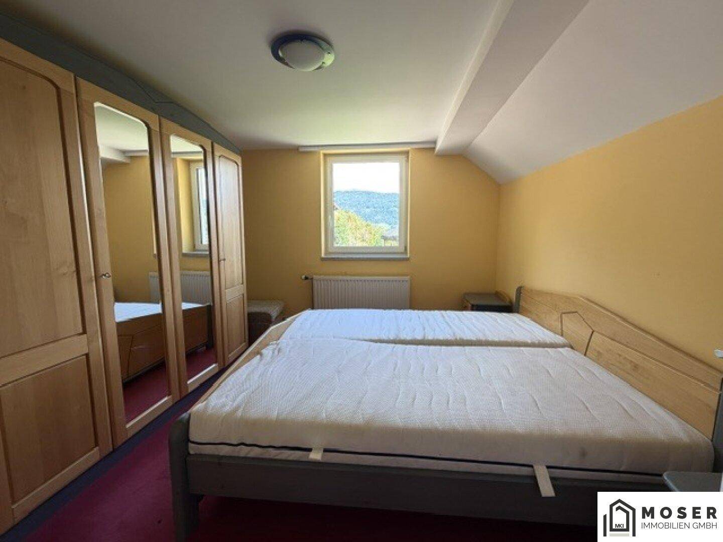 Schlafzimmer mit Doppelbett, Holzschrank und rotem Teppichboden unter Dachschrägen, mit Ausblick.