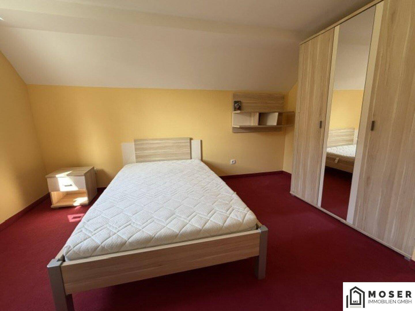 Schlafzimmer mit Einzelbett, Holzschrank mit Spiegeltür und rotem Teppichboden, unter Dachschräge.