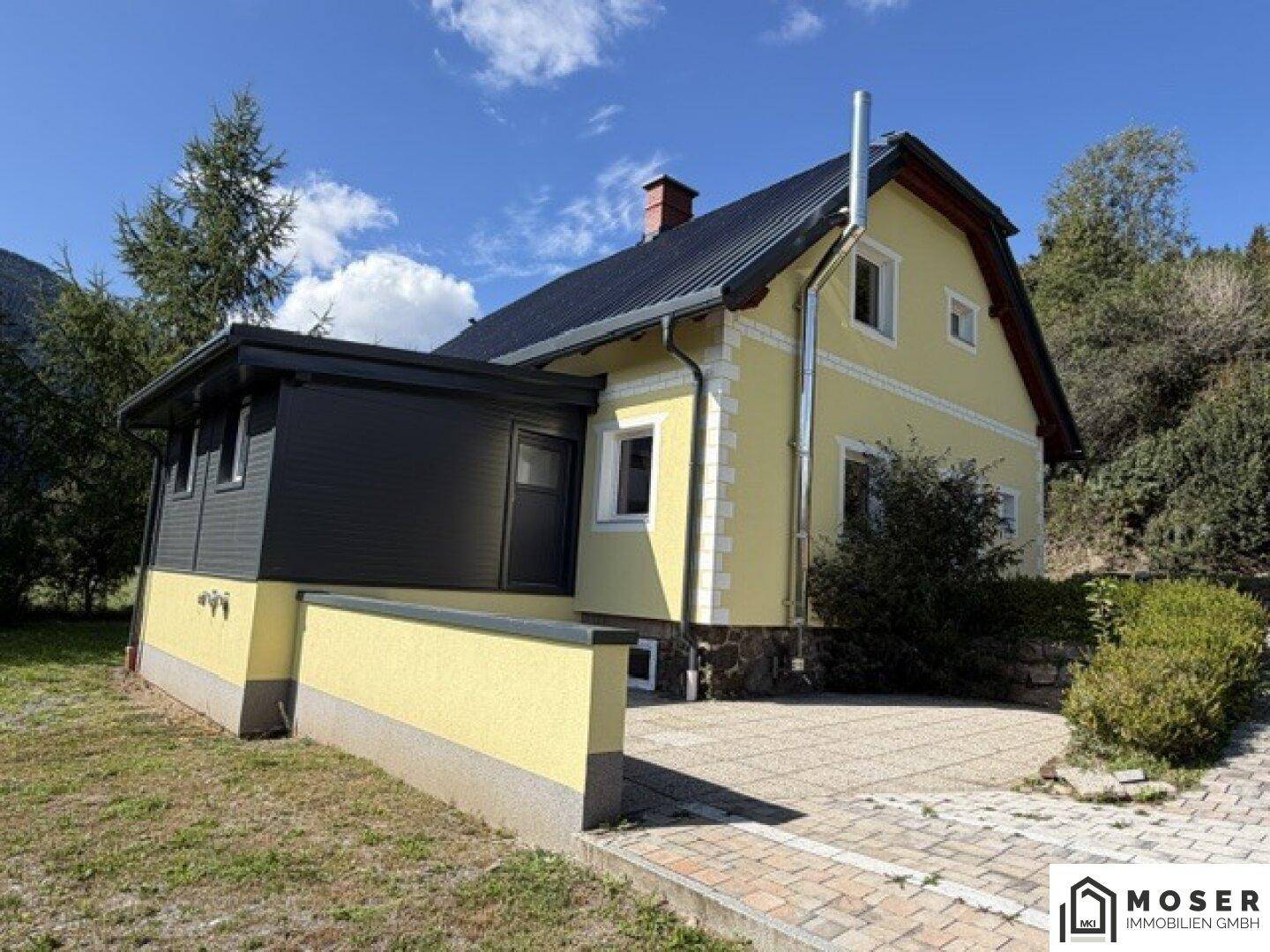 Seitenansicht des gelben Hauses mit gepflastertem Bereich und Blick auf die umliegende Landschaft.