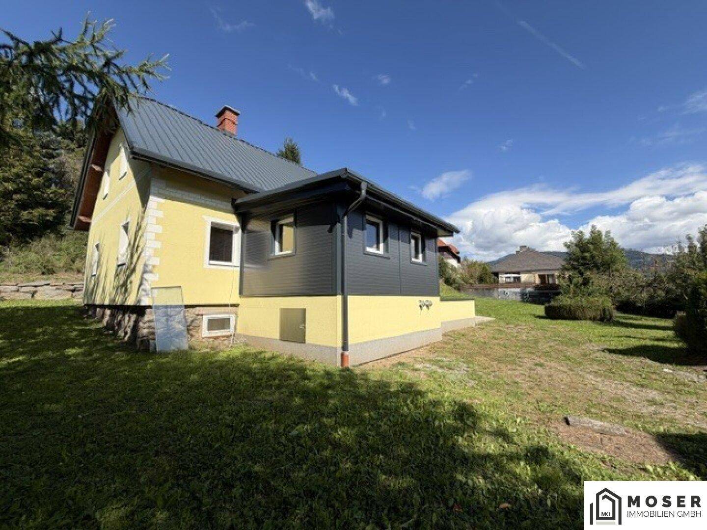 Das gelbe Haus mit dunkelgrauem Anbau, umgeben von einer weitläufigen grünen Rasenfläche.