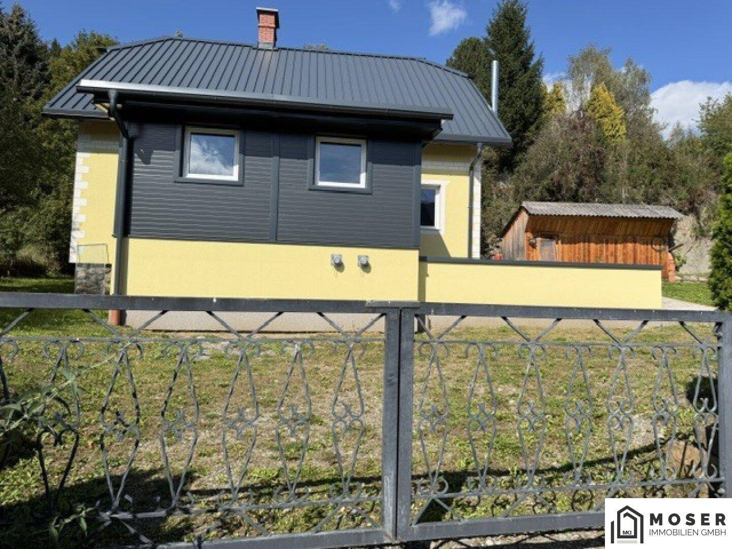 Gelbes Haus mit dunkelgrauem Anbau, umgeben von Grünflächen und einem schmiedeeisernen Zaun.