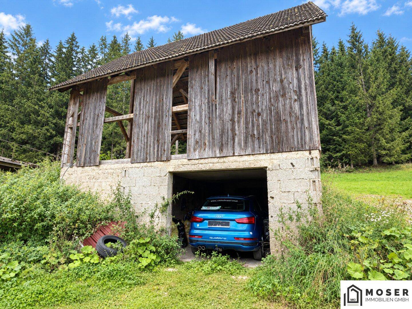 Altes Holzgebäude mit integrierter Garage, in der ein blaues Auto parkt, umgeben von Wald.