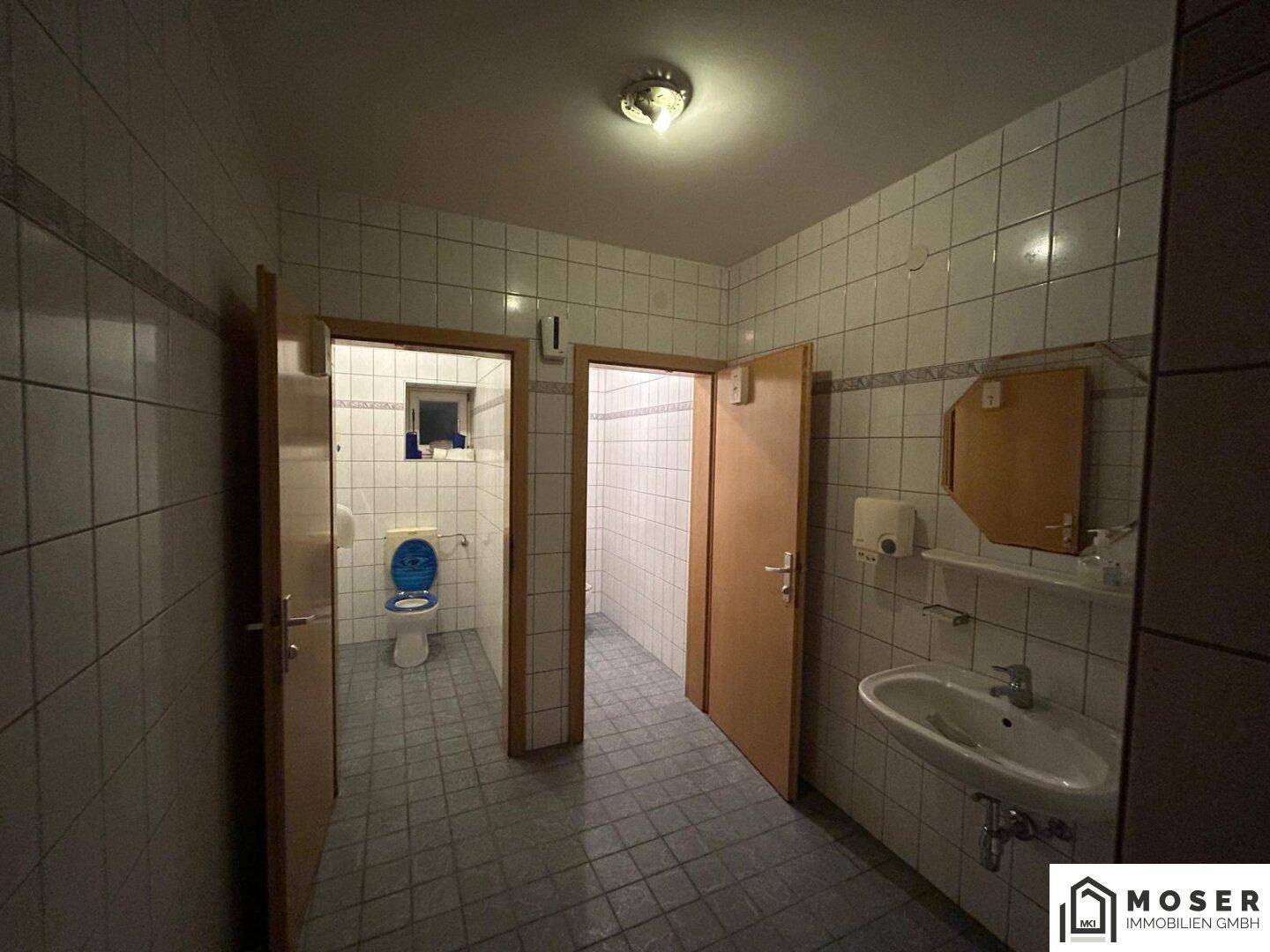 Toilettenbereich mit Fliesenboden, Waschbecken und zwei Holztüren zu den Kabinen.