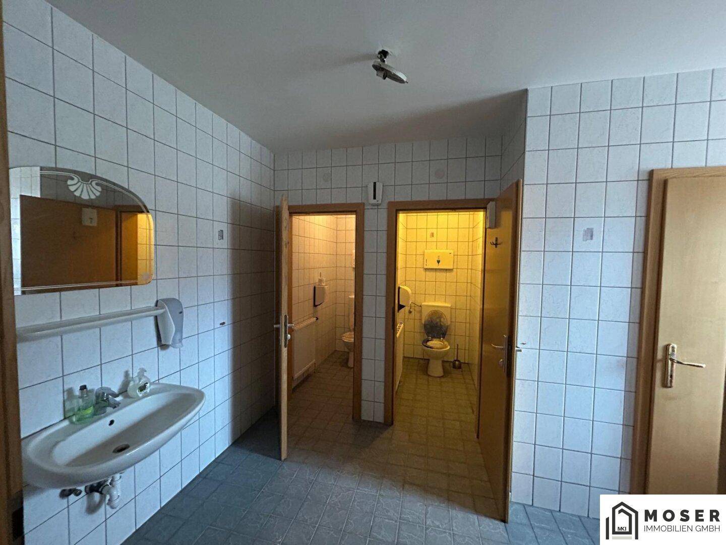 Toilettenbereich mit Fliesenboden, Waschbecken und Spiegel, sowie zwei Holztüren.