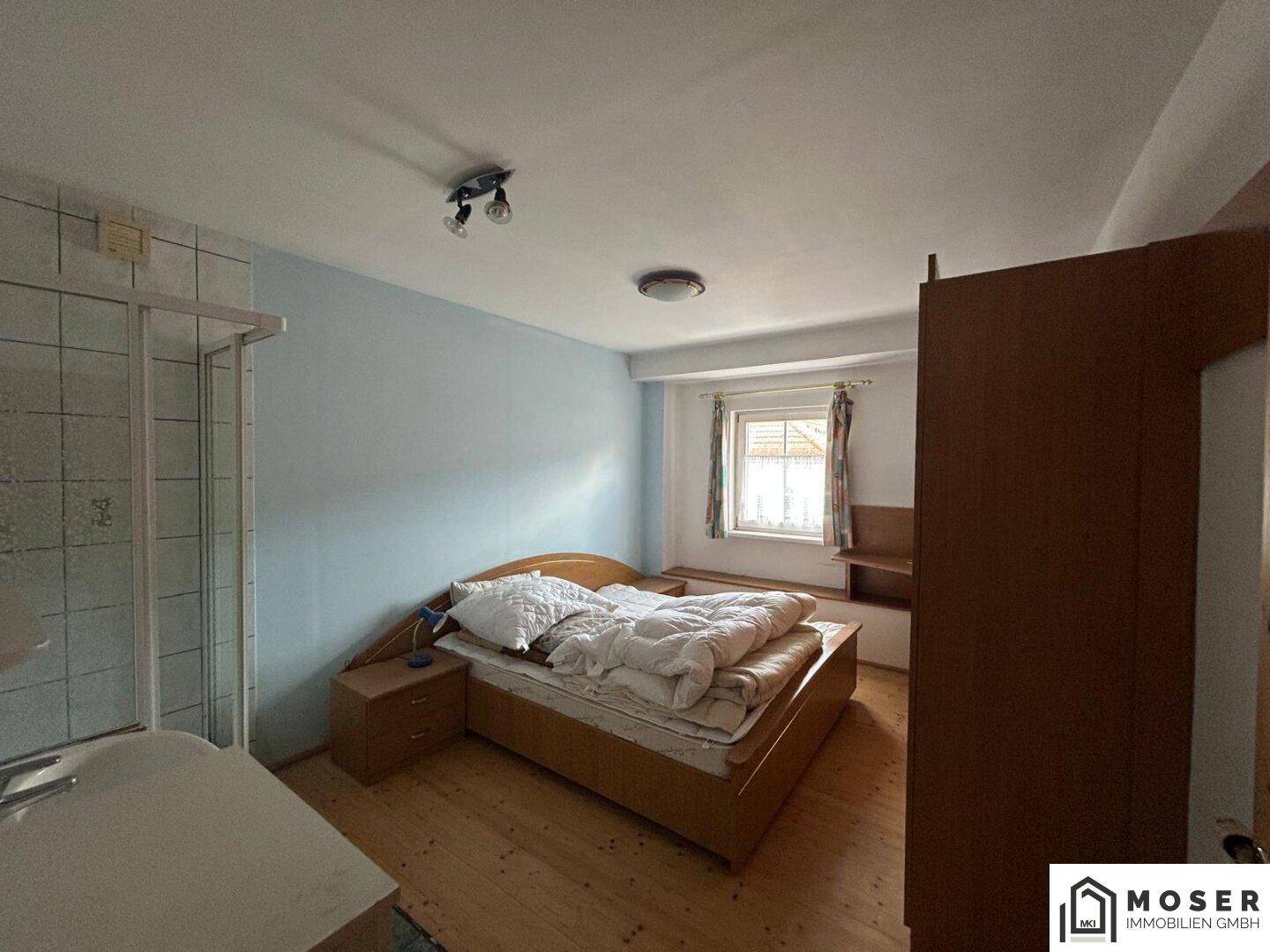 Schlafzimmer mit hellblauer Wand, Holzdielenboden, Bett und einer Duschkabine im Raum.