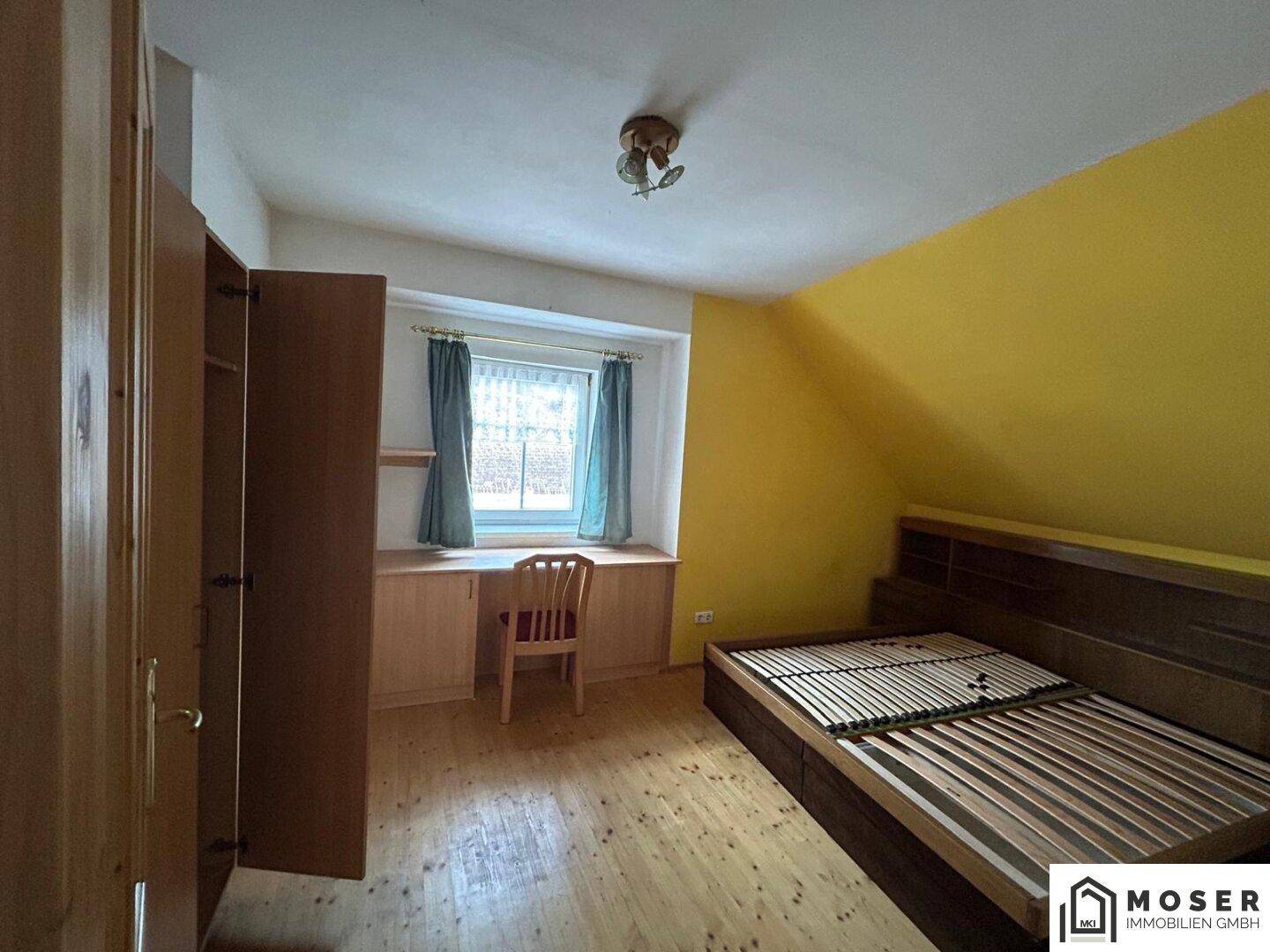 Schlafzimmer mit gelber Wand, Holzdielenboden, Bettgestell und einem Schreibtisch am Fenster.