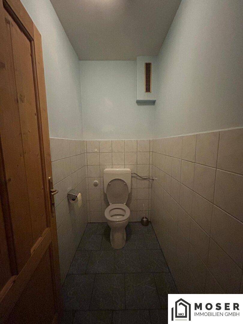 Separate Toilette mit Fliesenboden, halbhohen Fliesenwänden und einem Fenster.