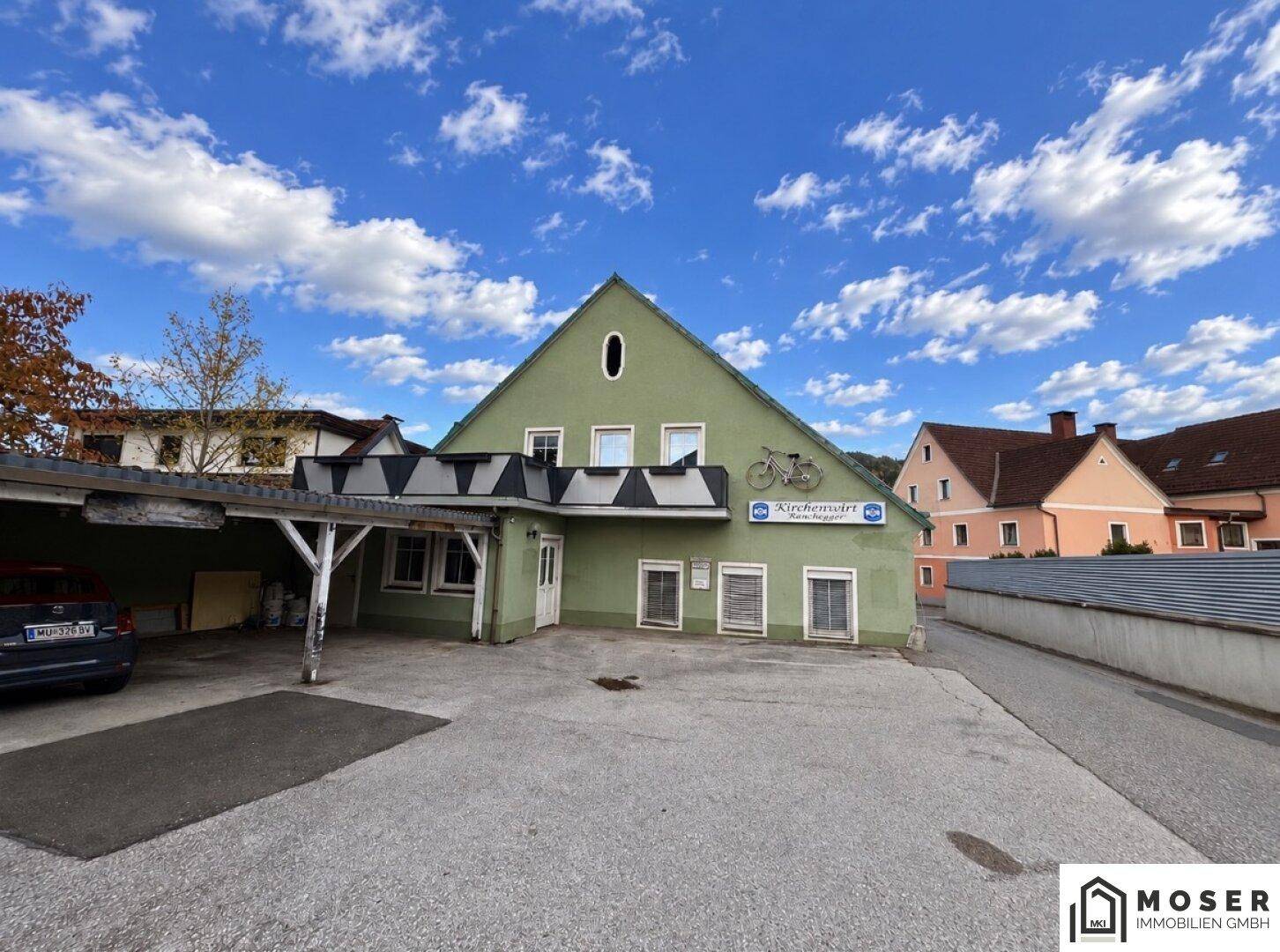 Grünes Gebäude mit Giebeldach, Carport und großem Parkplatz davor unter blauem Himmel.