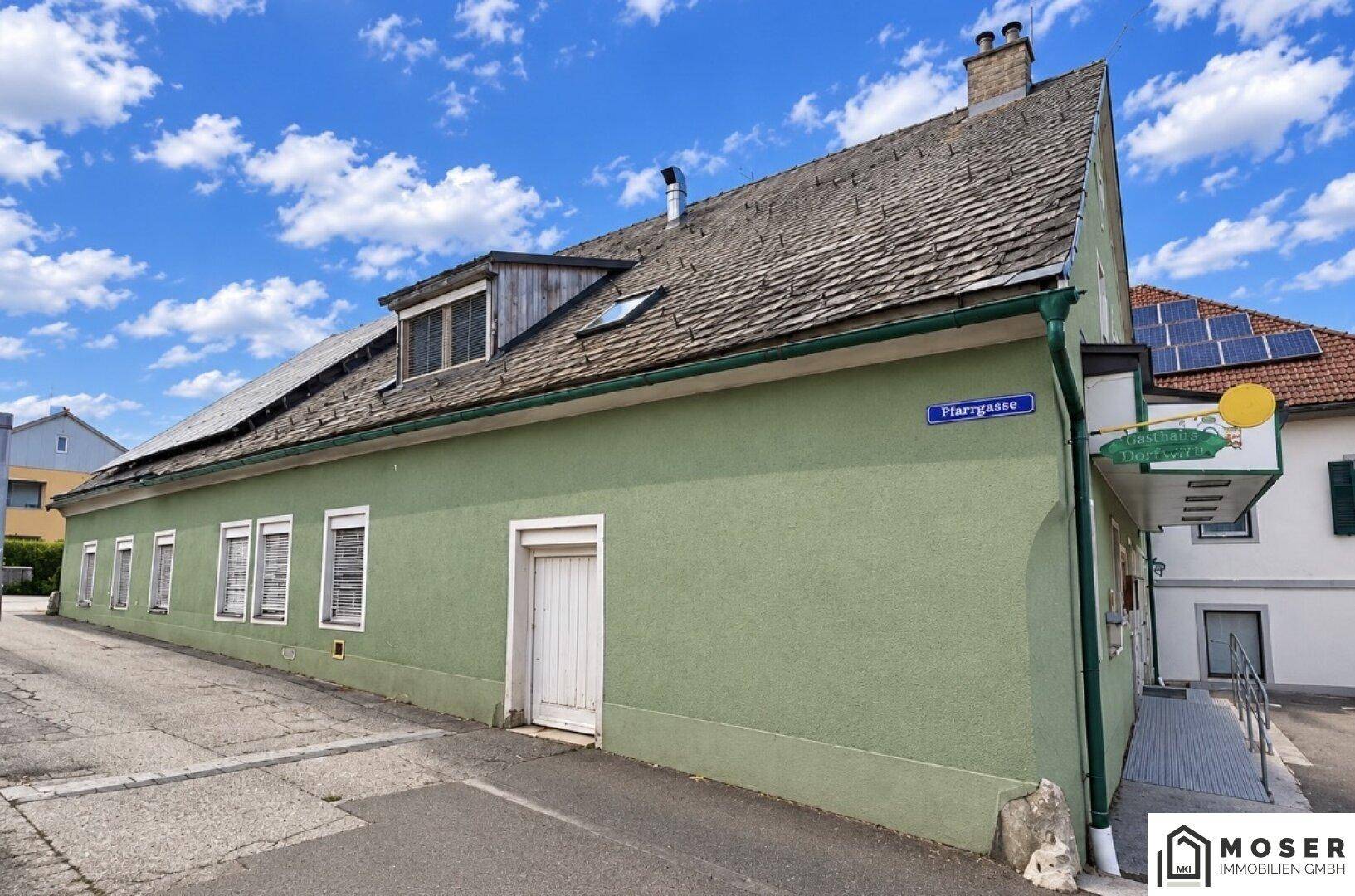 Seitenansicht eines grünen Gebäudes mit altem Dach und einer Gasse davor unter blauem Himmel.
