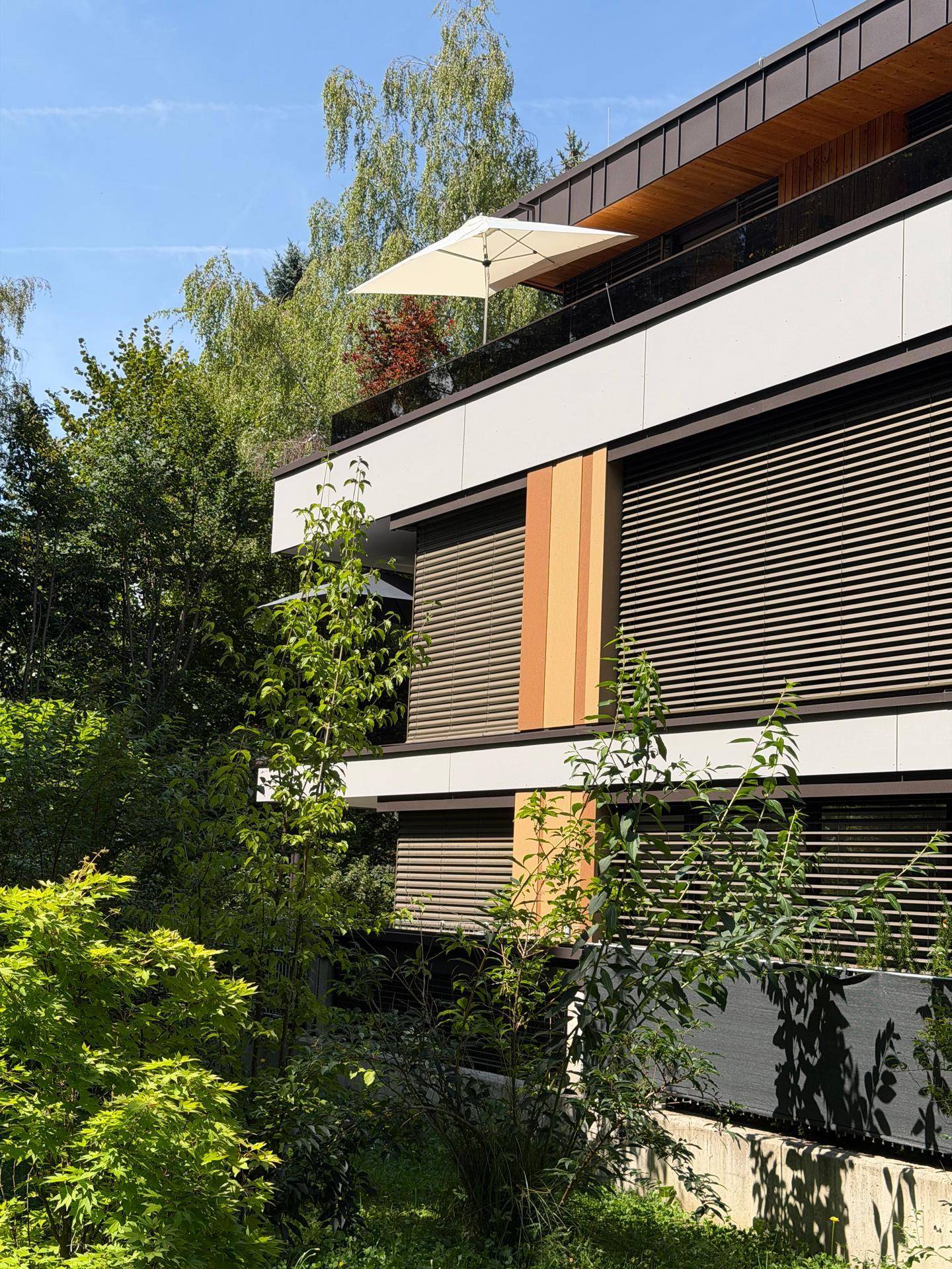 Moderne Gebäudefassade mit Holz- und Weißtönen, horizontalen Jalousien und einem Sonnenschirm auf der oberen Terrasse.