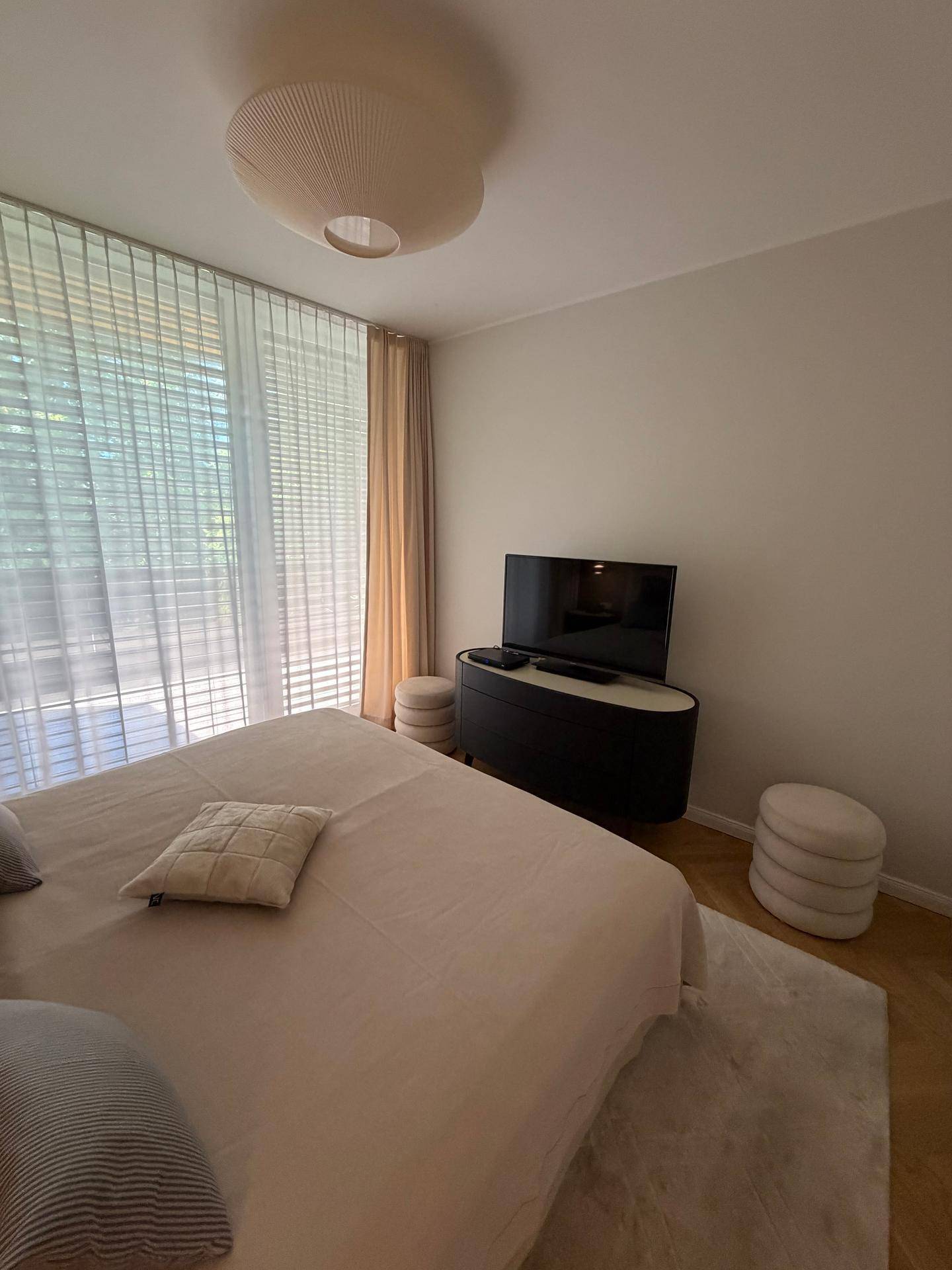 Helles Schlafzimmer mit großem Bett, Fernseher und bodentiefen Fenstern, die viel Licht hereinlassen.