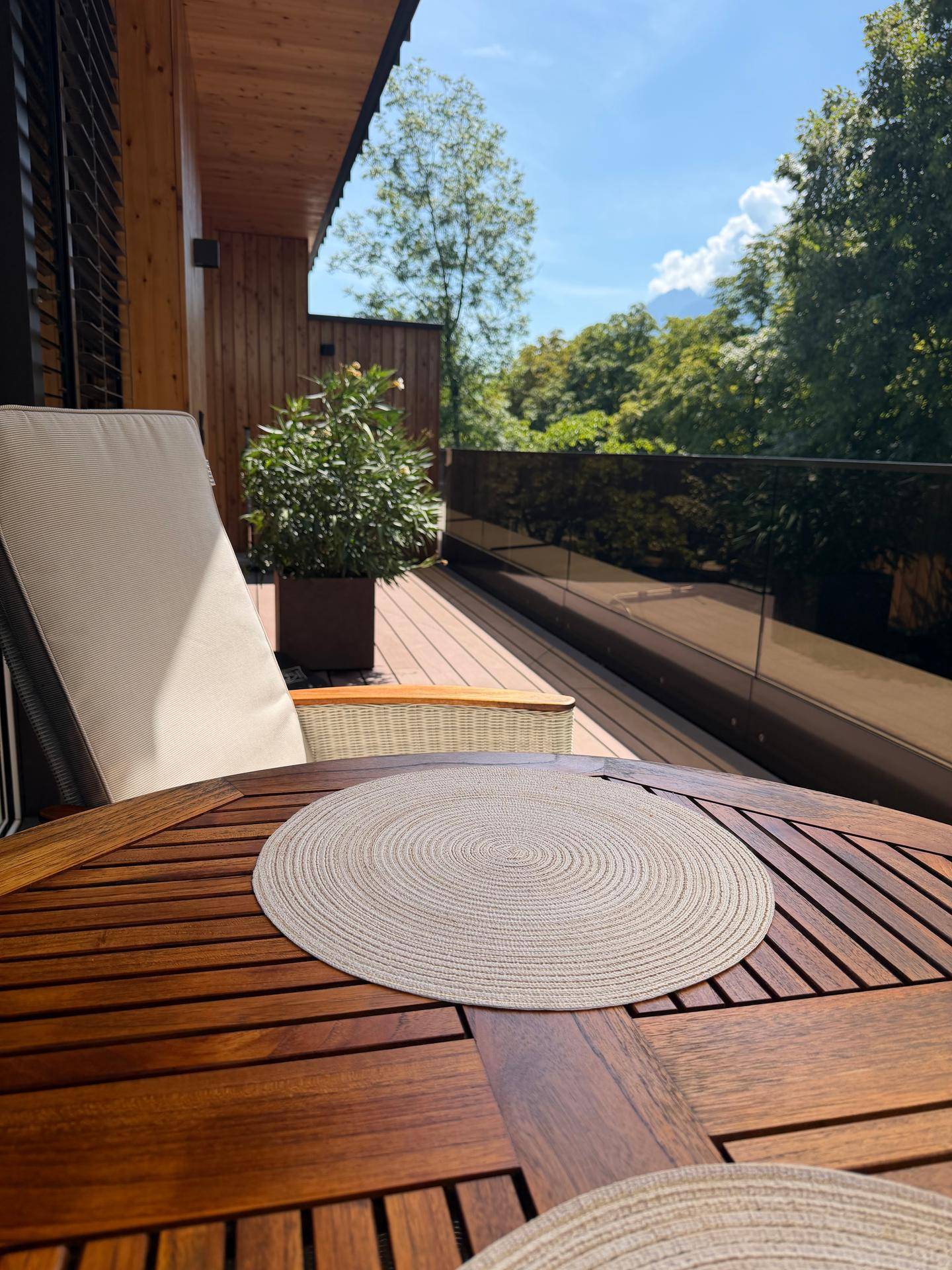 Gemütlicher Außenbereich mit Holztisch, Liegestuhl und Pflanzen auf einer sonnigen Terrasse mit Bergblick.