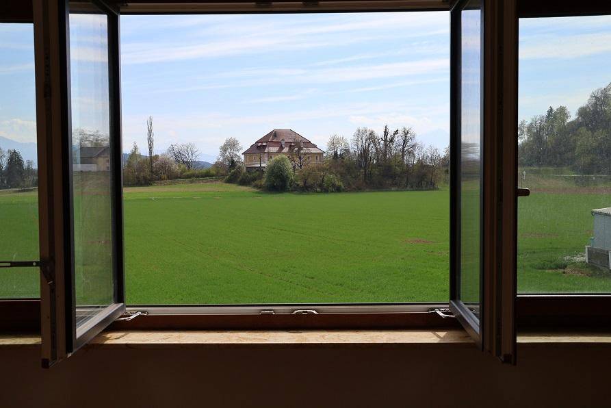 Weitblick aus einem Fenster über grüne Felder zu einem entfernten Gebäude und Bergen.