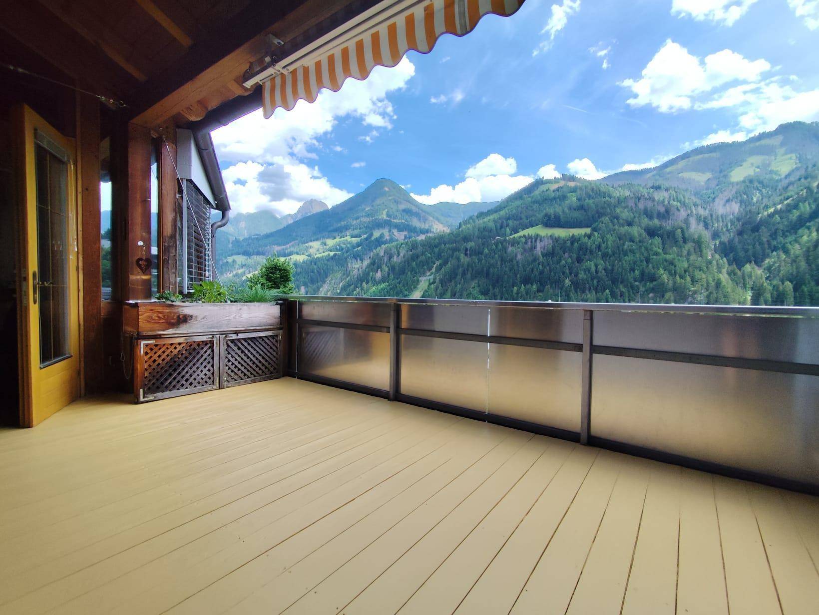 Geräumiger Balkon mit Holzboden, Markise und atemberaubendem Bergblick auf die grüne Landschaft.