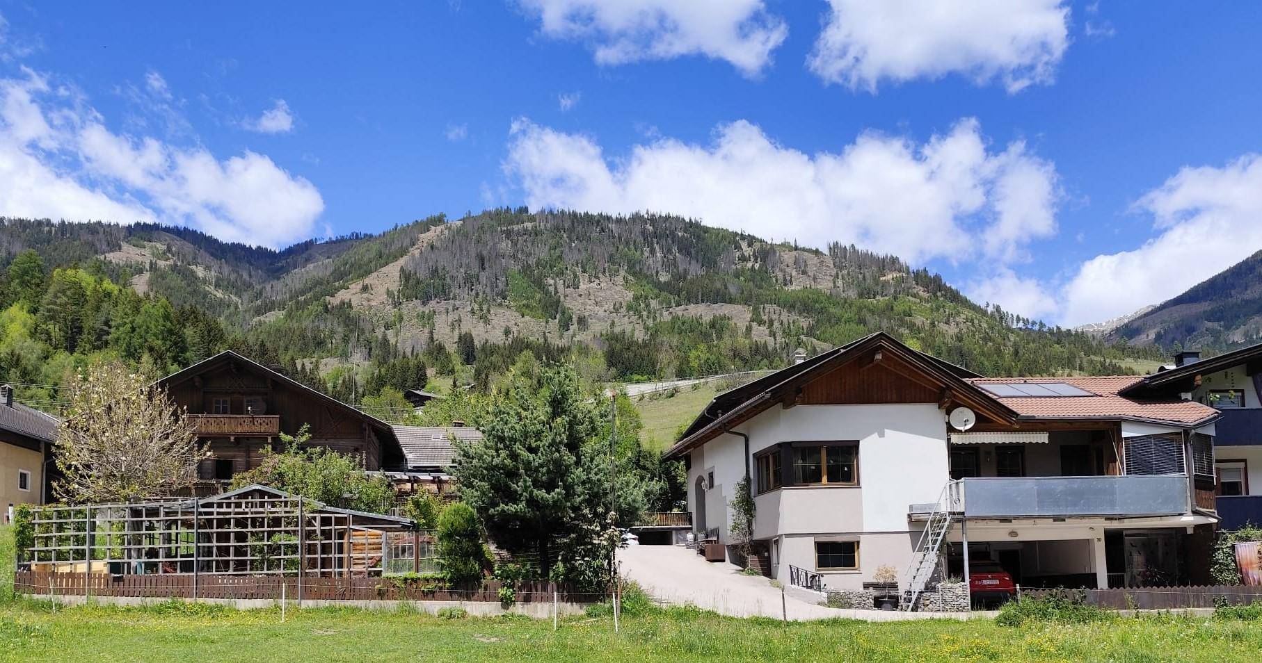 Haus in alpiner Landschaft mit grünen Bergen und weitem blauem Himmel.