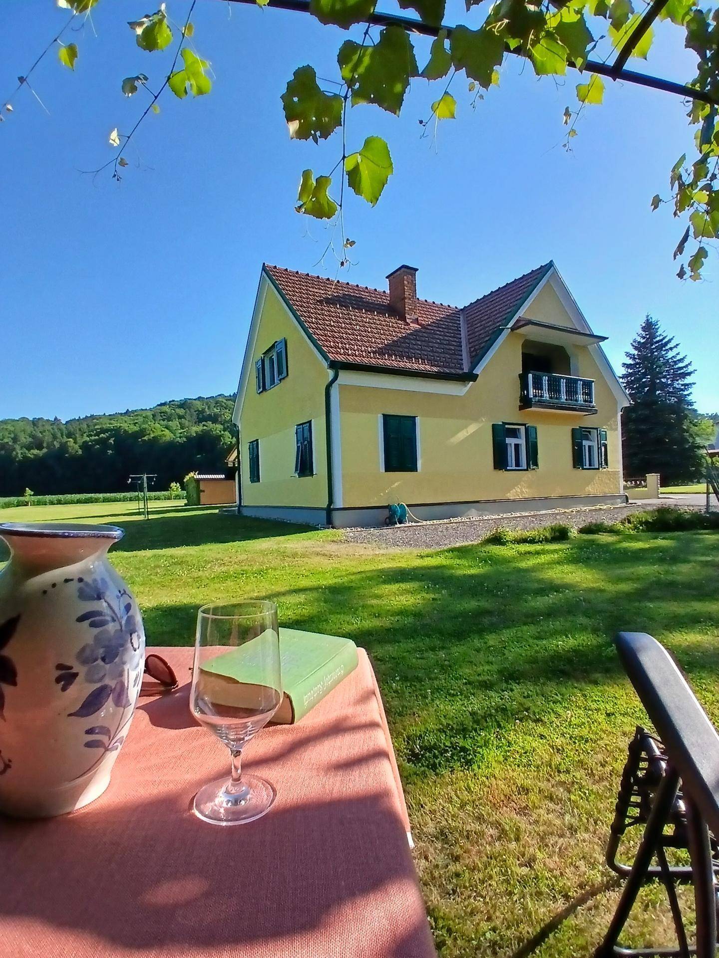 Idyllisches gelbes Haus mit rotem Dach, umgeben von grünen Wiesen und Bäumen unter blauem Himmel.