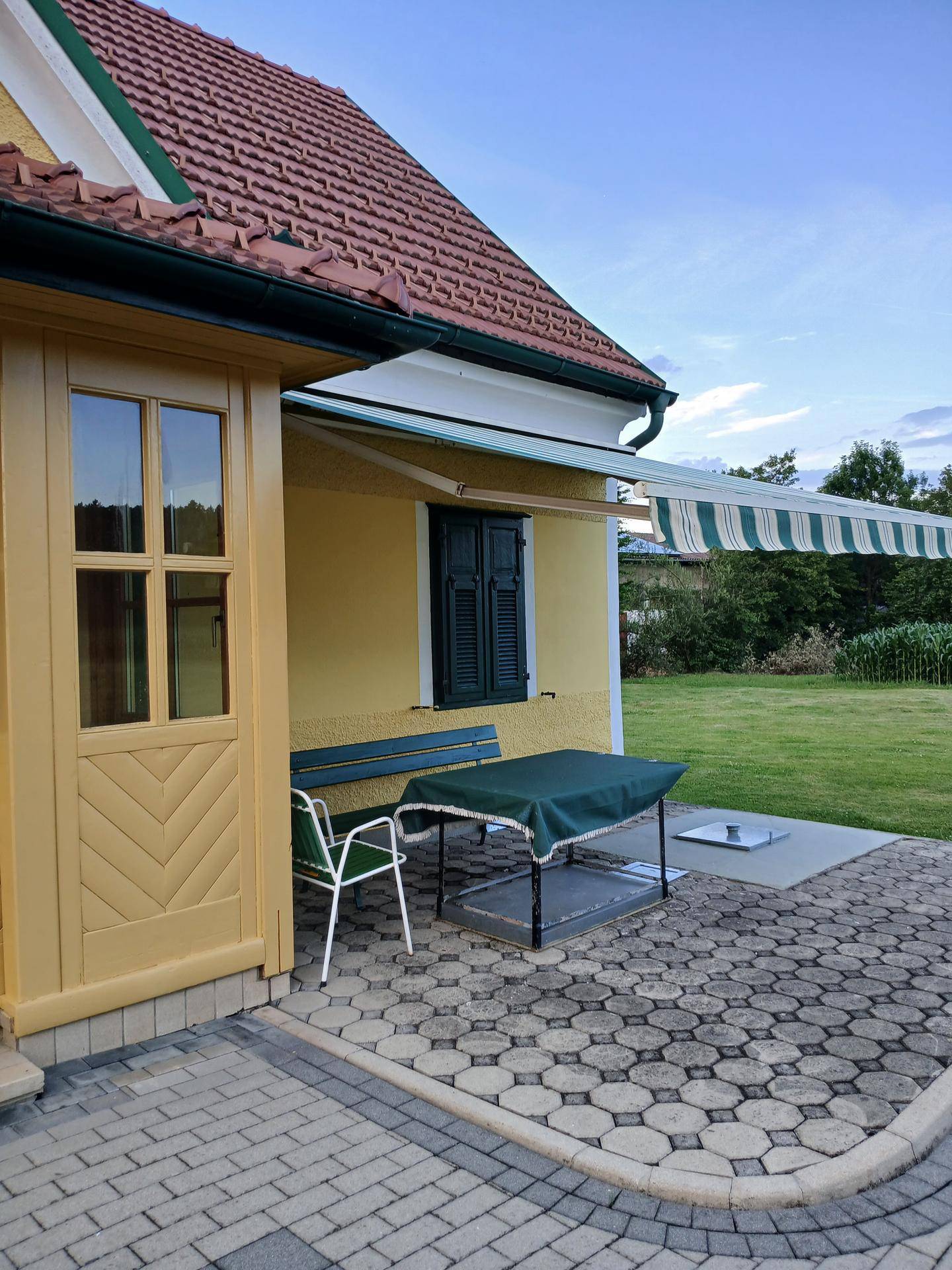 Gepflasterte Terrasse neben dem Haus mit Markise, Gartenbank und Blick auf den Rasen.
